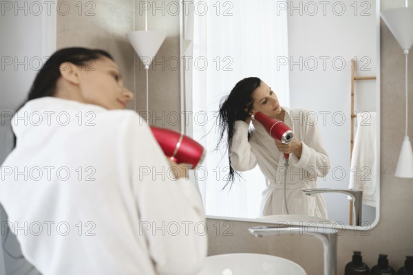 In a stylish bathroom, a woman in a white robe uses a hairdryer to dry her wet hair. Sunlight