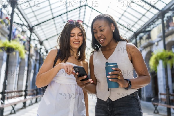 Two happy businesswomen walking through a city gallery, sipping coffee and using a smartphone,