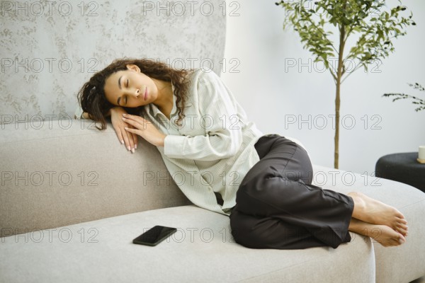 A young woman is comfortably curled up on a light-colored couch, peacefully napping. Soft sunlight