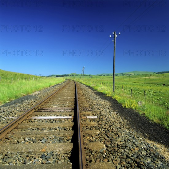 The railway track stretches through a lush green rural area, flanked by vibrant grasslands and a