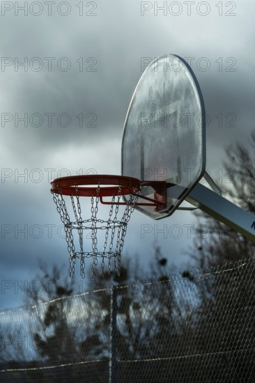 A solitary basketball hoop stands under overcast skies as dusk approaches. The chain net hangs