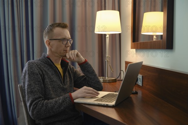 Middle-aged man sits at a wooden desk in a hotel room, focused on his laptop and is watching a