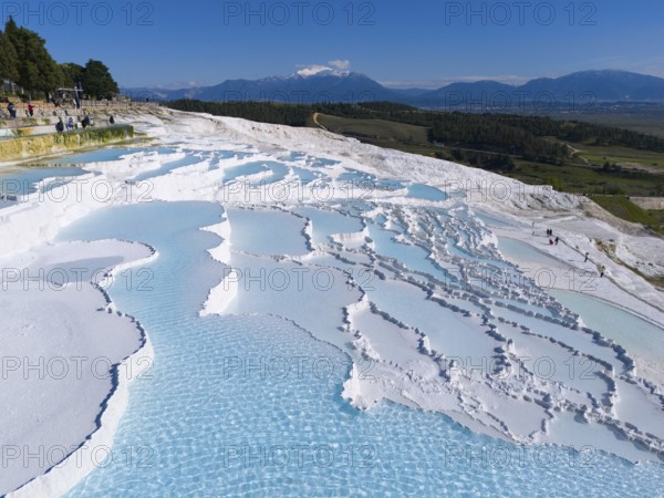 Wide view over white cascade terraces with turquoise blue water against a mountain backdrop, aerial