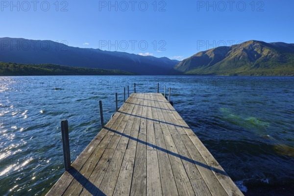 A narrow footbridge leads into the blue waters of a lake, surrounded by an impressive mountain