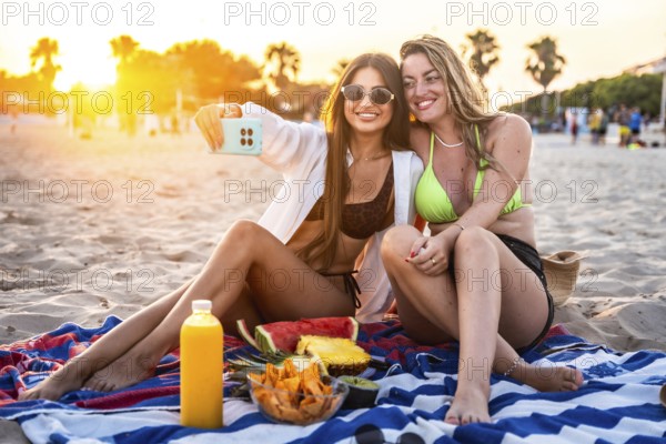 Two female friends are enjoying a beach picnic at sunset, taking a selfie with fresh fruit and
