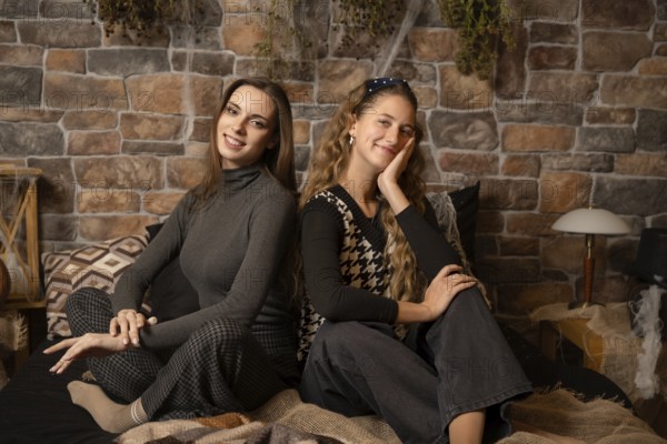 Two young women pose comfortably in a cozy indoor space featuring a rustic stone wall. The