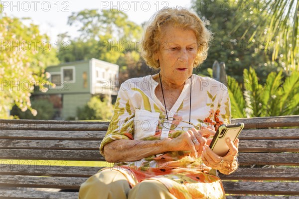 Senior woman sitting on a wooden bench in a sunny park, using a mobile phone to stay connected