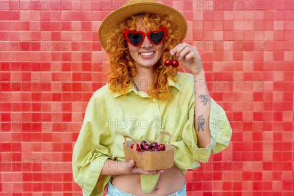 Happy young woman with curly red hair eating cherries from a small cardboard box, wearing heart