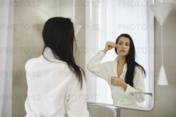 A woman dressed in a white robe stands before a mirror, carefully applying moisturizing serum in a