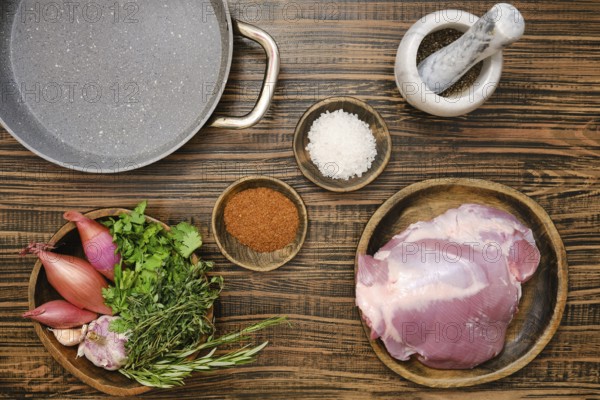 Fresh turkey thigh on a wooden plate alongside vibrant herbs, shallots, and spices. Overhead view