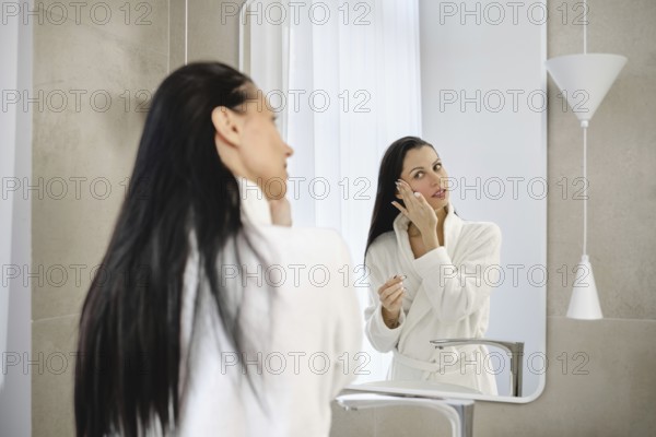 In a stylish bathroom, a woman in a white robe washing off makeup in front of the mirror. She