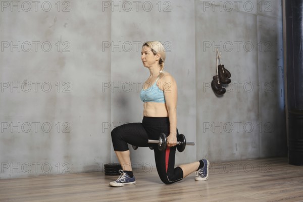 A woman with blonde hair demonstrates proper form while doing lunges with a dumbbell. She exercises