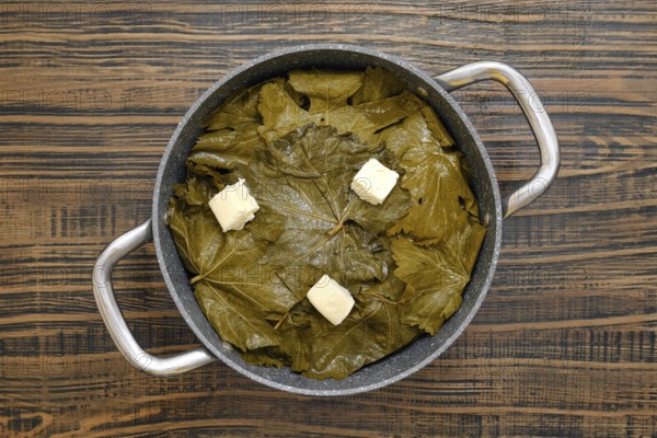 A pot filled with grape leaves prepares to cook dolma on a wooden table. Butter sits on top, ready