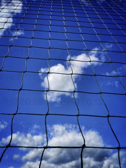 Looking up at a blue sky filled with clouds, framed by a black net. The sunlight highlights the