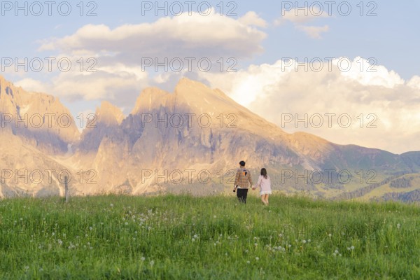 A couple walks across a flowering meadow towards the mountain range at sunset, Alpe di Siusi,
