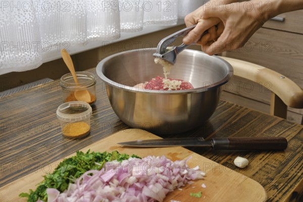 Hands mincing garlic into a metal bowl filled with ground meat while chopped onions and fresh herbs