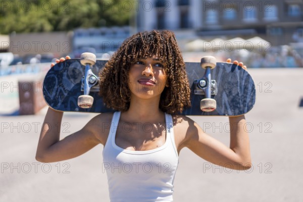 Confident young woman holding skateboard on shoulders, enjoying sunny day at skatepark, showcasing