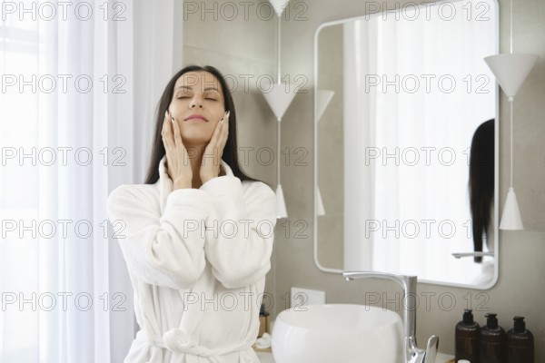 A woman in a white robe enjoys a moment of self-care in a stylish bathroom. She applies skincare