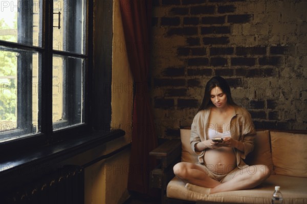 A woman in her third trimester sits comfortably on a sofa at home. She is wearing casual clothing