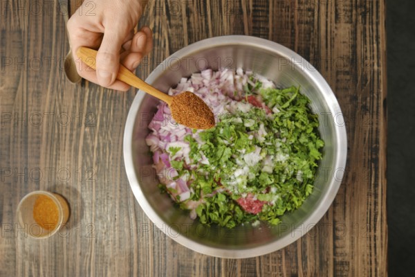 A hand holds a wooden spoon filled with spices over a stainless steel bowl containing chopped