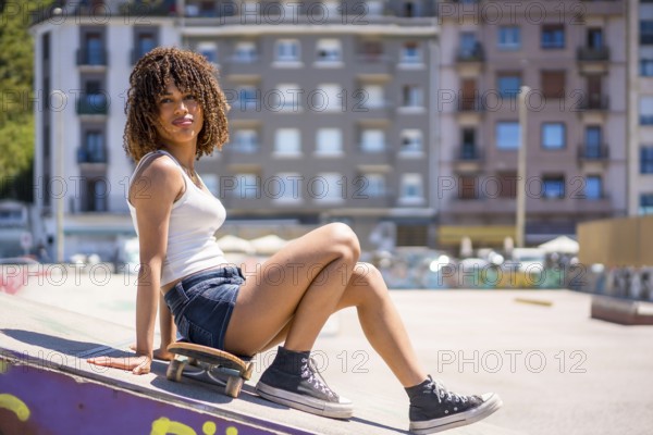 Young woman sitting on her skateboard atop a ramp in an urban skatepark, soaking up the sun on a