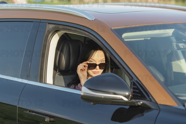 A woman with long dark hair wears stylish sunglasses while sitting in a sleek black car. The sun