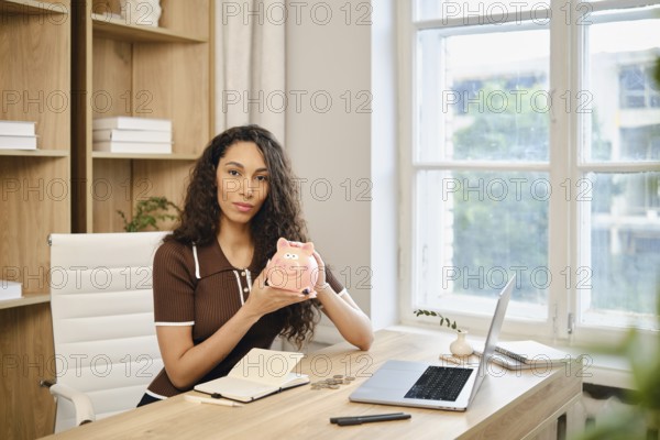 A woman with long curly hair sits at a desk in a bright home office, holding a piggy bank. She is