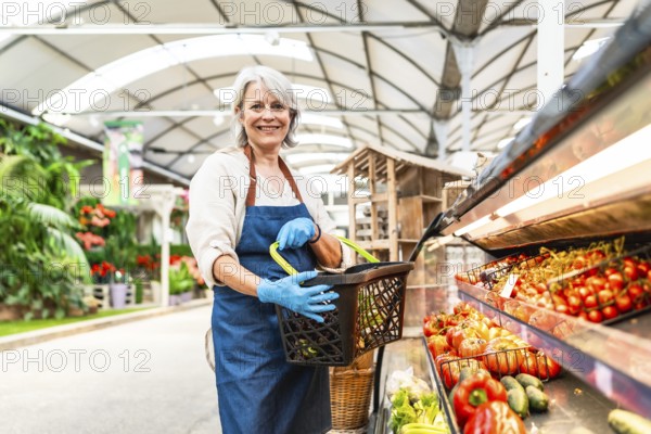 Senior greengrocer wearing gloves holding a shopping basket and smiling in a supermarket with fresh