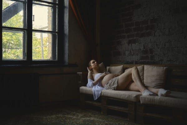 A pregnant woman lounges comfortably on a cozy couch in her home. The warm sunlight filters through