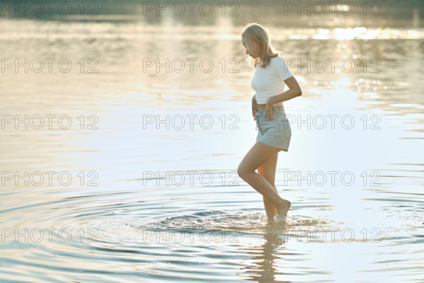 A woman is walking barefoot in gently rippling water at a tranquil lake during sunset. The warm