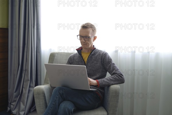 A man wearing glasses sits comfortably in a gray chair and fills out a tax return online on his