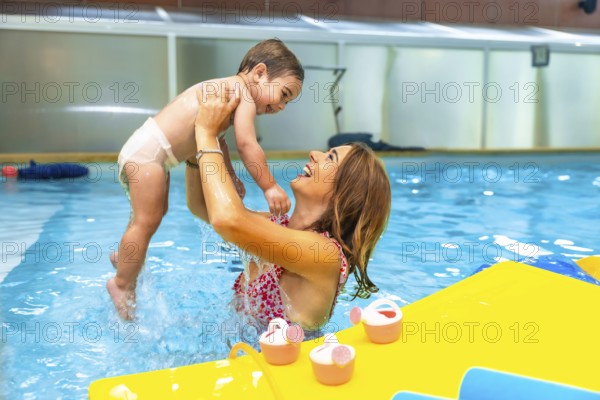 Mother playfully lifting smiling baby out of water in indoor swimming pool during matinee swim