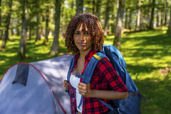 Young woman wearing a backpack enjoys camping in a serene forest, relaxing near her tent and
