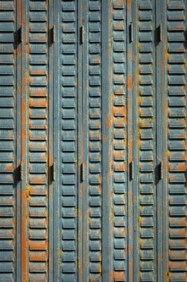 A close-up view of weathered metal shutters displaying a mix of blue paint and rust. The surface