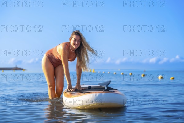 Smiling woman paddle surfing in calm blue waters, enjoying a sunny summer day. Clear sky and