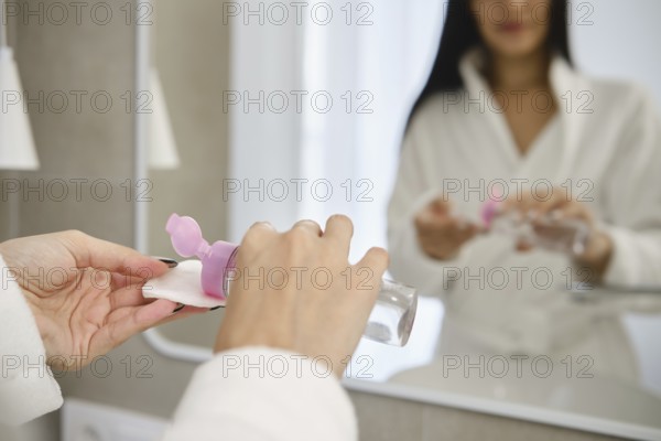 Reflection of a woman applying micellar water to a cotton sponge pad in a bright bathroom during