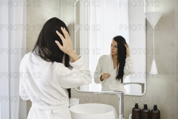 A woman stands in front of a mirror in a modern bathroom, dressed in a white bathrobe. She is