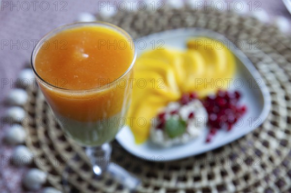A glass with multi-coloured juice and a plate with mango slices and pomegranate seeds, Lower