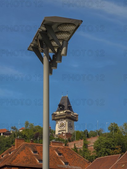 Clock tower behind a street lamp, surrounded by historic roofs and trees Graz Clock Tower Graz