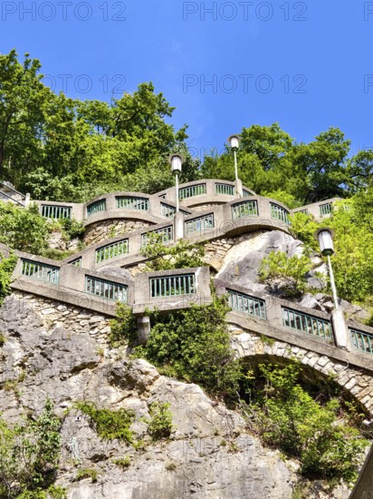 Stone wall with green railings and lamps on a steep, wooded slope under a blue sky Graz Styria