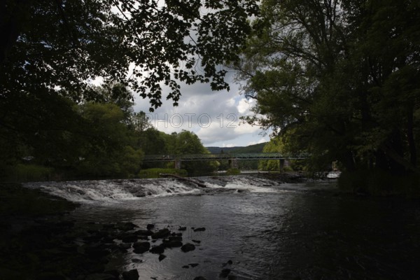 A dark river with a waterfall and bridge, surrounded by forests and cloudy skies Hardegg