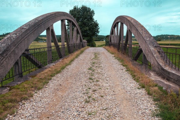 An old bridge with a gravel path leads through a rural area, surrounded by fields and a clear sky