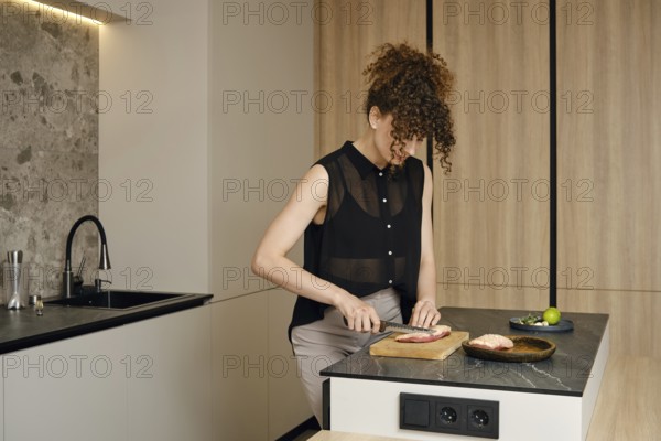 A woman with curly hair is cutting raw meat in a kitchen. Natural light illuminates her workspace