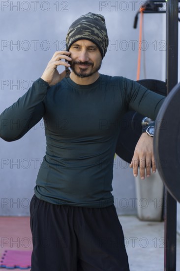 Athlete takes a break from his workout, leaning against a weightlifting rack and talking on his