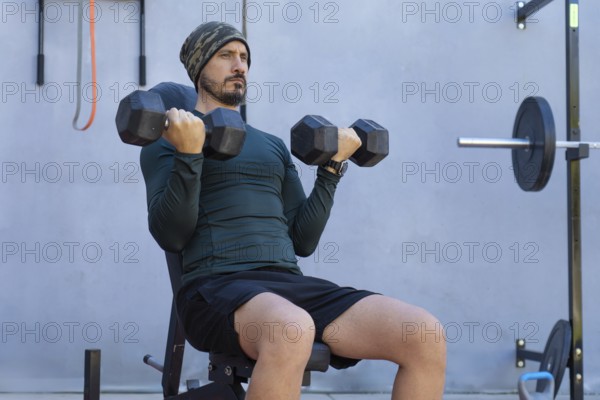 Focused male athlete sitting on a weight bench, lifting dumbbells and performing bicep curls during