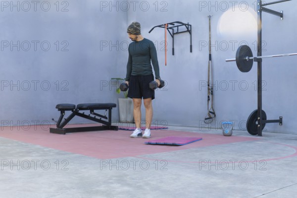 Young sportsman lifting two black dumbbells while engaging in a workout in a private outdoor gym,