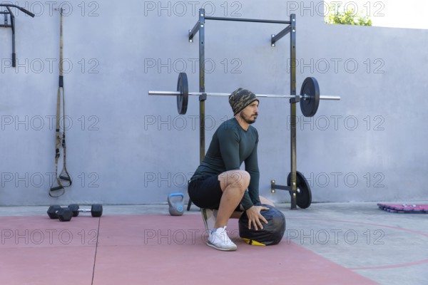 Young sportsman wearing beanie and thermal shirt performing wall ball squats in an outdoor gym,