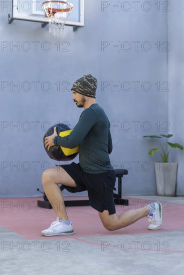 Young sportsman wearing a beanie and sportswear is lunging with a medicine ball in an outdoor gym,