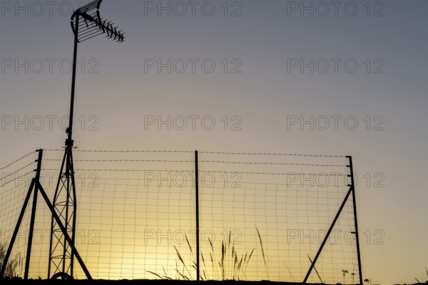 Silhouette of a television antenna and a wire mesh fence against a vibrant sunset sky