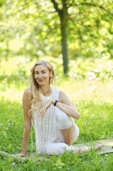 A woman dressed in white sits gracefully on a mat in a lush park, surrounded by vibrant green grass